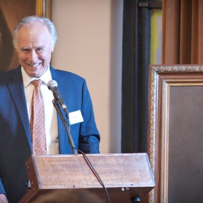 John Alexander standing at a podium smiling at an audience. To his left is a painting of a solemn, bearded, Henry George.