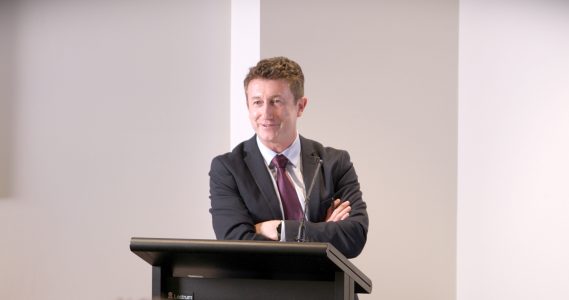 A man standing at a lecturn with his arms crossed a slight smile on his face.